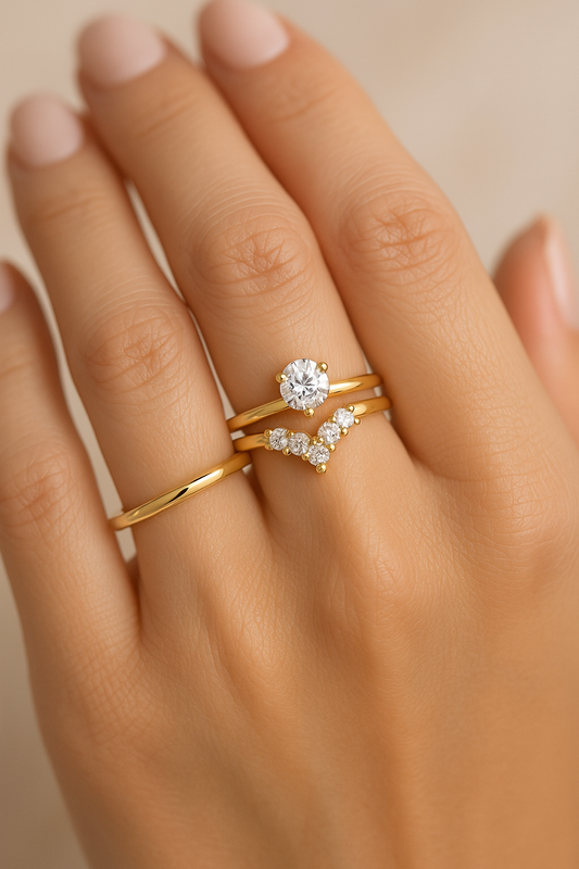 A woman’s hand wearing a diamond solitaire engagement ring stacked closely with a 14K gold graduated V-shaped diamond band, shown on a natural model hand for a delicate, elegant stacked look