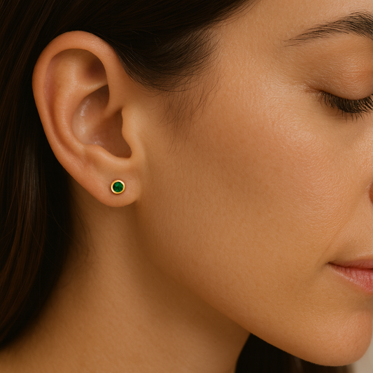 Close-up of a woman's ear wearing a small, round 3 MM emerald stud earring in a polished yellow gold bezel setting, with smooth skin and dark hair softly framing the face.